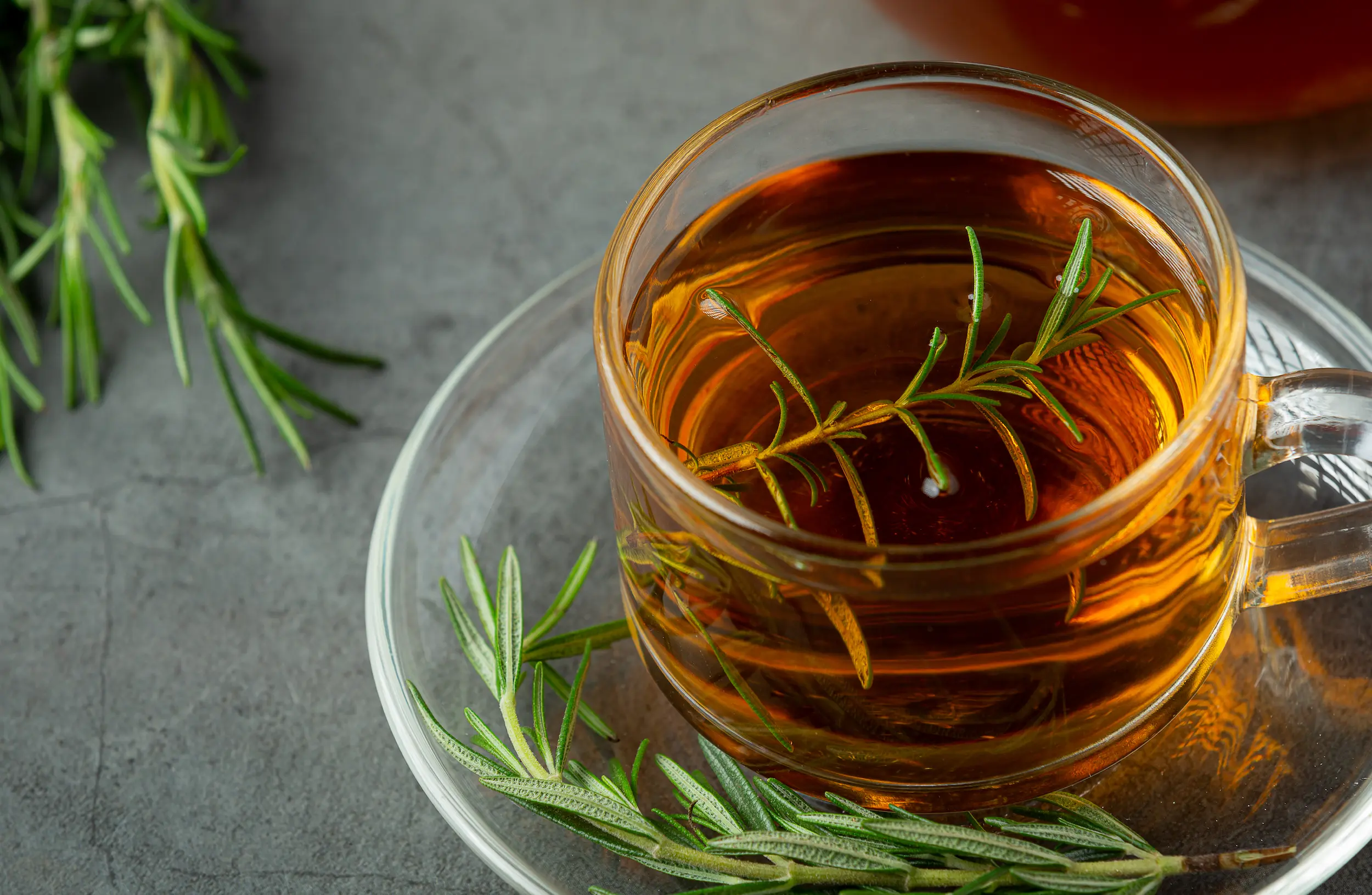 A clear glass mug of amber rosemary tea with fresh rosemary sprigs, on a dark textured surface